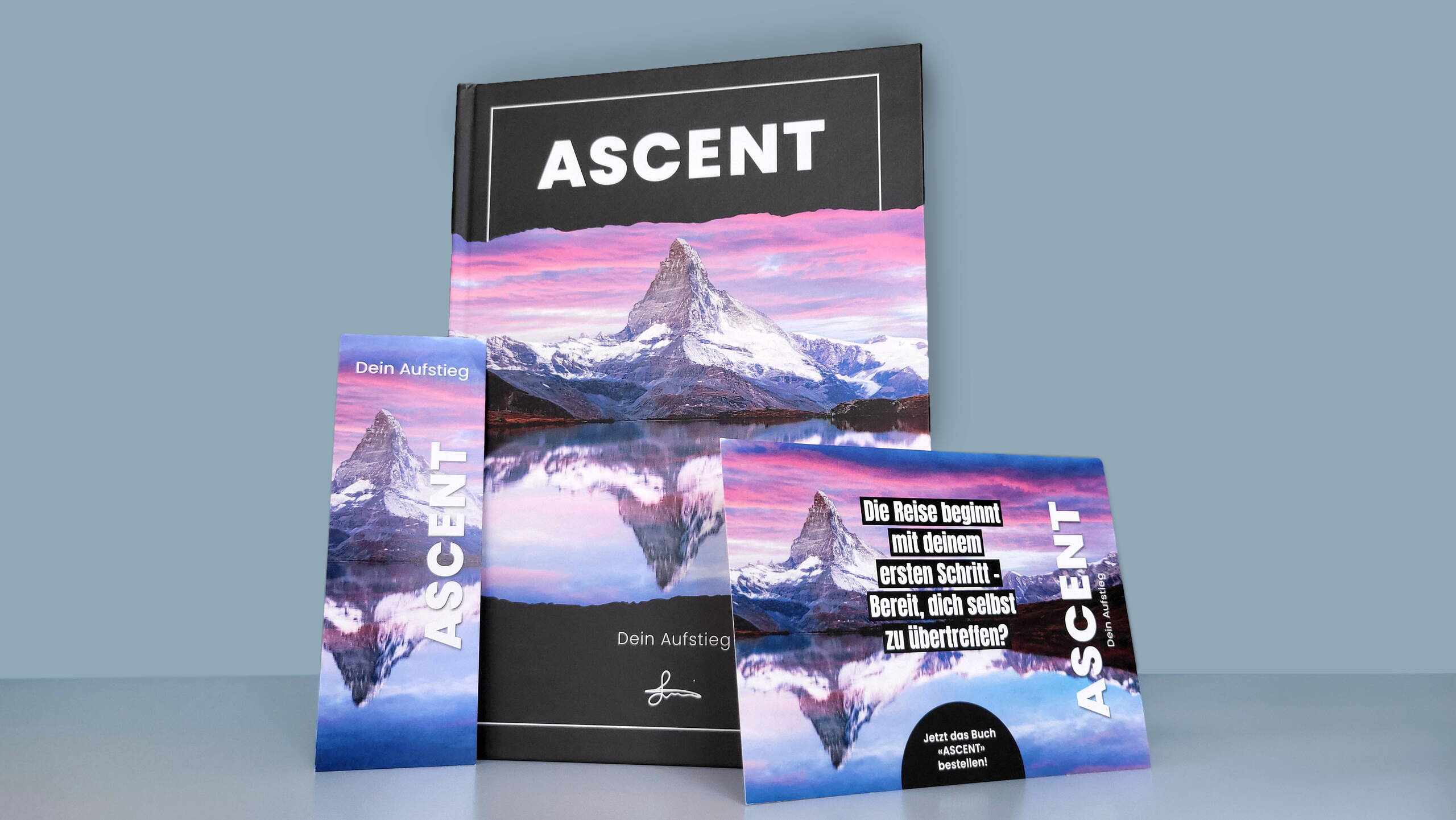 Vor einem grauen Hintergrund stehen das Buch, das Lesezeichen sowie die Bestellkarte von ASCENT – Dein Aufstieg